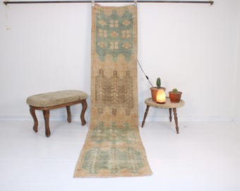 May include: A long, rectangular rug with a beige background and a repeating pattern of green and brown geometric shapes. The rug is hanging from a rod and is displayed on a white wall. A small wooden stool and a table with a small lamp are in the foreground.