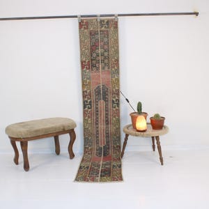 Peut inclure: Un tapis kilim turc vintage avec des motifs géométriques dans des tons de rouge, bleu et beige est suspendu à une tringle contre un mur blanc. Le tapis est présenté avec un petit tabouret en bois et des cactus en pot.