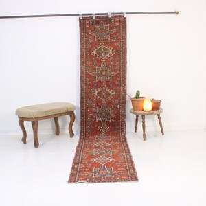 May include: A long, rectangular, vintage rug with a red and brown geometric pattern. The rug is hanging from a metal rod. A small, wooden stool with a beige cushion and a small table with plants and a lamp are also in the image.