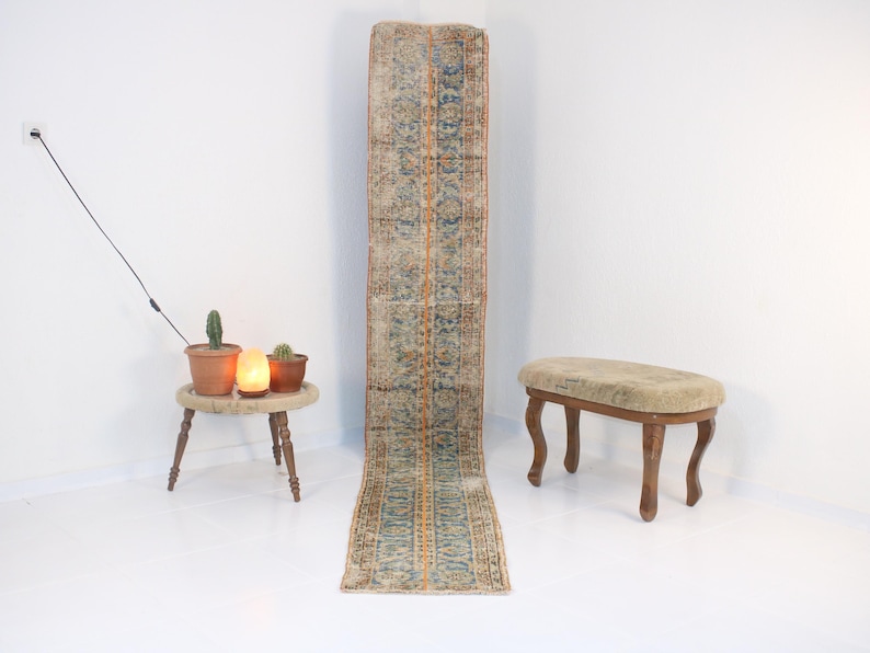 May include: A long, rectangular rug with a blue and orange pattern. The rug is on a white floor in front of a white wall. There is a small wooden stool with a beige cushion on the floor to the right of the rug.