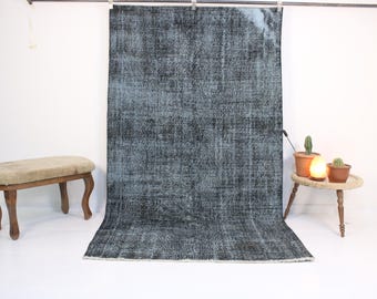 May include: A large, rectangular rug with a distressed, vintage appearance. The rug features a blend of dark gray and light blue hues, creating a textured, faded effect. The rug is displayed against a white backdrop, showcasing its design and color variations.