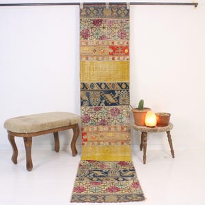 May include: A long, rectangular rug with a patchwork design. The rug features a variety of colours, including yellow, red, blue, and pink. The rug is hanging vertically against a white wall. A small, round table and a bench are in the foreground.