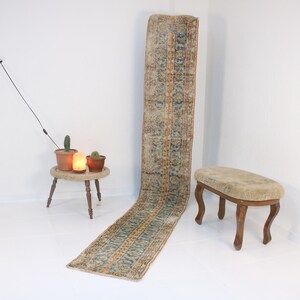 May include: A long, rectangular rug with a blue, orange, and beige pattern. The rug is laying on a white floor. A small wooden stool with a beige cushion is in the foreground.