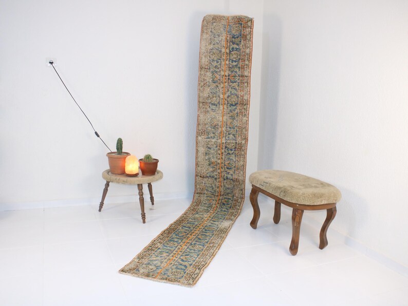 May include: A long, rectangular rug with a blue, orange, and beige pattern. The rug is laying on a white floor. A small wooden stool with a beige cushion is in the foreground.