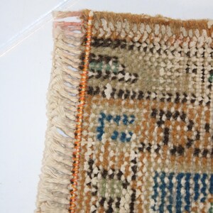 May include: Close-up of the fringe edge of a woven rug with a geometric pattern in shades of brown, blue, green, and white. The fringe is white with an orange thread running along the edge.