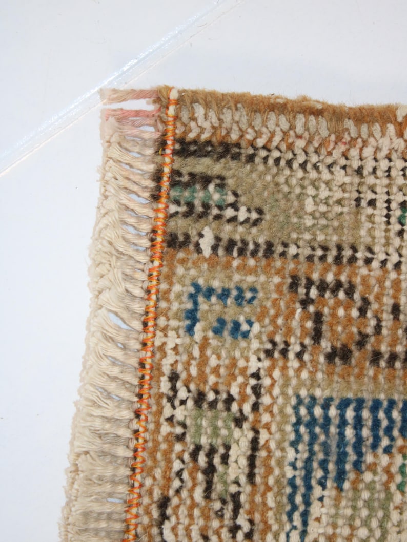 May include: Close-up of the fringe edge of a woven rug with a geometric pattern in shades of brown, blue, green, and white. The fringe is white with an orange thread running along the edge.