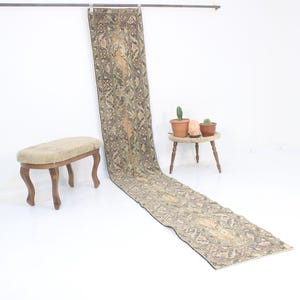 Könnte beinhalten: Ein langer, rechteckiger Teppich mit einem beige, grünen und braunen Muster. Der Teppich hängt an einer Metallstange und liegt auf einem weißen Boden. Ein kleiner Holzhocker mit einem beigefarbenen Kissen befindet sich im Vordergrund.