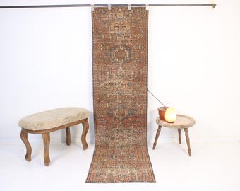 May include: A long, rectangular, vintage rug with a detailed geometric pattern in shades of red, brown, and blue. The rug is hanging vertically against a white wall. A small, round table and a wooden bench are in the foreground.