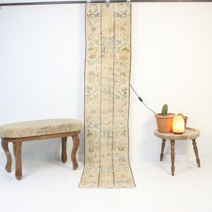 Könnte beinhalten: Ein langer, schmaler Vintage-Teppich mit floralem Muster in Beige, Blau und Rosa hängt an einer Stange. Rechts steht ein kleiner, runder Tisch mit einer Lampe und Topfpflanzen. Links befindet sich eine Holzbank mit gepolsterter Oberseite.