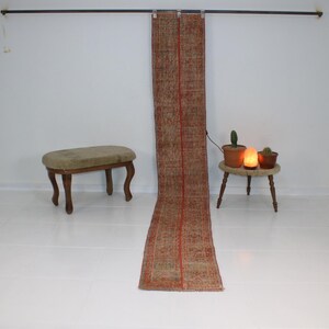 May include: A long, narrow, red and brown patterned rug hangs from a rod against a white wall. A small green plant sits on a wooden stool next to the rug.