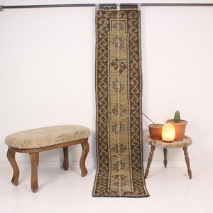 May include: A long, rectangular, brown and beige rug with geometric patterns hangs against a white wall. A small, round, wooden stool with a light and a cactus sits to the right. A wooden bench with curved legs is in the foreground.