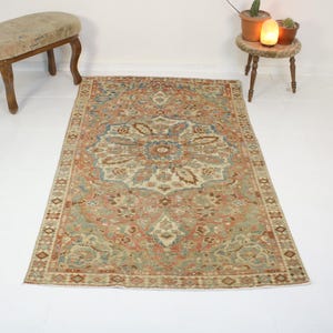 May include: A rectangular, vintage-style rug with an intricate floral pattern in shades of red, blue, beige, and green. The rug is placed on a white surface, showcasing its detailed design and aged appearance.