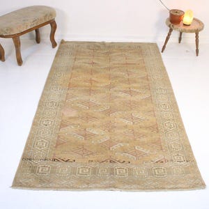 May include: A rectangular area rug featuring a muted tan and beige color palette with a repeating geometric diamond pattern across the center field and a wide border decorated with a continuous Greek key motif in lighter cream and tan tones.