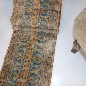 May include: A long, narrow rug with a repeating pattern of blue, orange, and green. The rug has a frayed edge and is made of a woven material.
