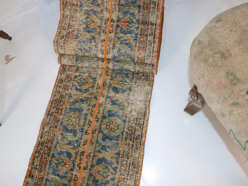May include: A long, narrow rug with a repeating pattern of blue, orange, and green. The rug has a frayed edge and is made of a woven material.