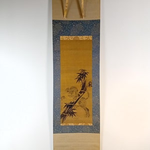 May include: A traditional Japanese scroll painting featuring a tiger and bamboo. The artwork is primarily in shades of gold, brown, and black, with a blue patterned border. The scroll is suspended by gold-colored ribbons.
