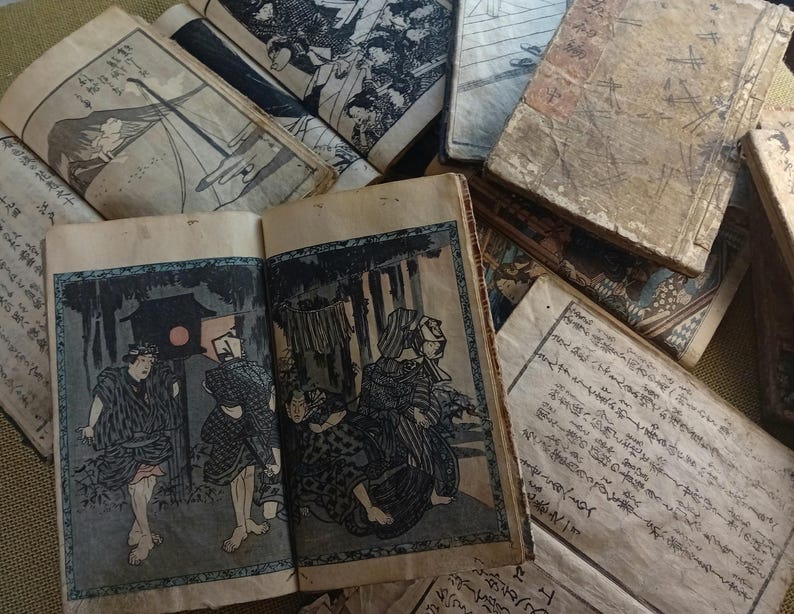 May include: A collection of antique Japanese woodblock print books. The open book displays a colorful illustration of figures in a dark setting. The covers and pages show signs of age, with faded colors and wear. The books are stacked together.