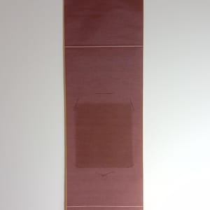 Op de afbeelding: Een bordeauxrode rol, waarschijnlijk voor kalligrafie of kunst, hangt verticaal tegen een witte muur. De rechthoekige rol heeft een lichtere rand en een opgerolde onderkant. Het heeft een centraal, lichter gekleurd vierkant met vage markeringen.