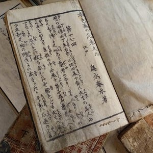 May include: An open antique book with aged, yellowed pages. The text is in Japanese calligraphy, with visible ink strokes. The book is part of a collection of old books, with visible wear and tear.