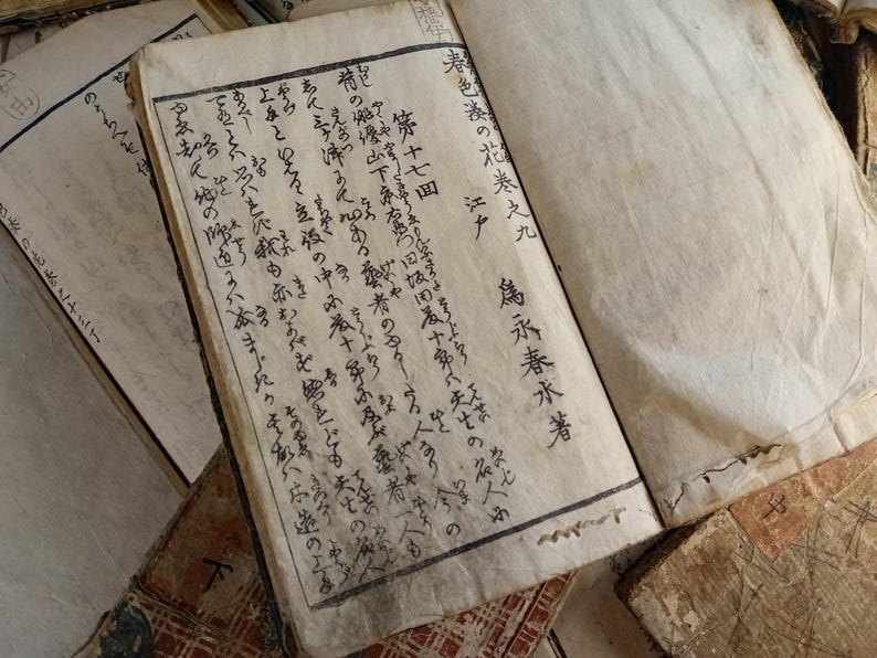 May include: An open antique book with aged, yellowed pages. The text is in Japanese calligraphy, with visible ink strokes. The book is part of a collection of old books, with visible wear and tear.