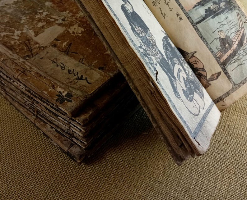 May include: Antique books with aged, textured pages. One book is open, revealing a detailed illustration in blue and black ink. The pages are yellowed with age, and the covers show signs of wear. The books are resting on a textured, brown surface.