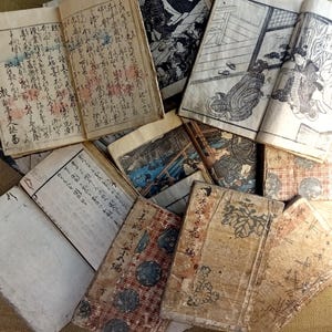 May include: A collection of antique Japanese books with aged, textured covers and pages. The books feature traditional illustrations and handwritten text in black ink. Some pages display colorful accents, and the overall aesthetic is vintage and historical.
