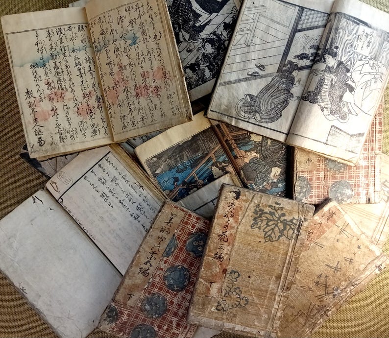 May include: A collection of antique Japanese books with aged, textured covers and pages. The books feature traditional illustrations and handwritten text in black ink. Some pages display colorful accents, and the overall aesthetic is vintage and historical.