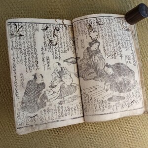 May include: An open antique book with Japanese text and illustrations. The pages feature detailed drawings of people in traditional clothing, with a dark wooden cylinder resting on the top right corner of the book.