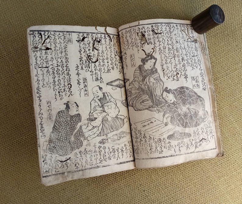 May include: An open antique book with Japanese text and illustrations. The pages feature detailed drawings of people in traditional clothing, with a dark wooden cylinder resting on the top right corner of the book.