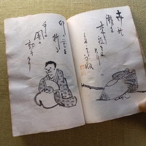 Puede incluir: Un libro antiguo abierto con caligraf&iacute;a japonesa manuscrita e ilustraciones a tinta. Una ilustraci&oacute;n representa a una persona con un abanico, la otra una figura de p&aacute;jaro. Una peque&ntilde;a piedra clara descansa en la p&aacute;gina derecha.
