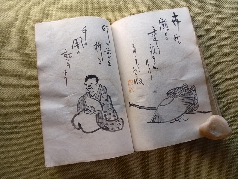 Puede incluir: Un libro antiguo abierto con caligraf&iacute;a japonesa manuscrita e ilustraciones a tinta. Una ilustraci&oacute;n representa a una persona con un abanico, la otra una figura de p&aacute;jaro. Una peque&ntilde;a piedra clara descansa en la p&aacute;gina derecha.