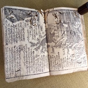 May include: An open antique book with detailed black and white illustrations and Japanese text. A dark wooden cylinder holds the book open. The pages depict scenes of people and landscapes.