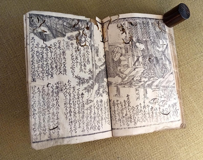 May include: An open antique book with detailed black and white illustrations and Japanese text. A dark wooden cylinder holds the book open. The pages depict scenes of people and landscapes.