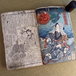 May include: An open antique book with Japanese text and illustrations. The right page features a colourful depiction of a person in traditional clothing near water, with a red orb in the background. A black cylindrical object is attached to the book.