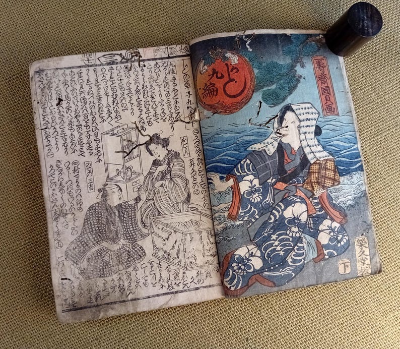 May include: An open antique book with Japanese text and illustrations. The right page features a colourful depiction of a person in traditional clothing near water, with a red orb in the background. A black cylindrical object is attached to the book.