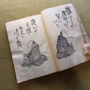 Puede incluir: Un libro abierto con caligraf&iacute;a japonesa e ilustraciones de figuras con vestimenta tradicional. Los dibujos son en tinta negra sobre papel envejecido. Una peque&ntilde;a piedra clara descansa en la p&aacute;gina derecha.