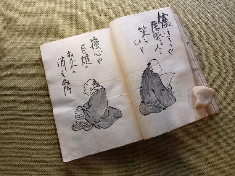 Puede incluir: Un libro abierto con caligraf&iacute;a japonesa e ilustraciones de figuras con vestimenta tradicional. Los dibujos son en tinta negra sobre papel envejecido. Una peque&ntilde;a piedra clara descansa en la p&aacute;gina derecha.