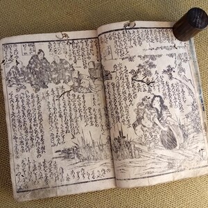 May include: An open antique book with Japanese text and illustrations. The pages are aged and show detailed black and white drawings of figures and landscapes. A dark wooden cylinder is attached to the top edge of the book.