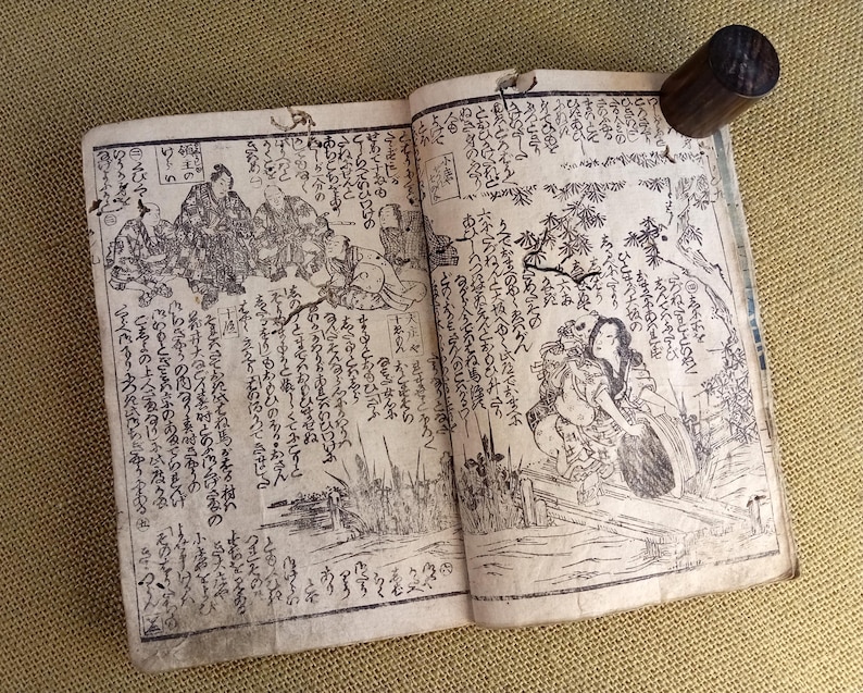 May include: An open antique book with Japanese text and illustrations. The pages are aged and show detailed black and white drawings of figures and landscapes. A dark wooden cylinder is attached to the top edge of the book.