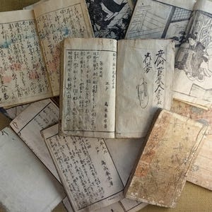 May include: A collection of antique Japanese books and documents, featuring handwritten text and illustrations. The pages are aged with visible wear and tear, showcasing the historical nature of the items. Some books are open, revealing detailed calligraphy and drawings.