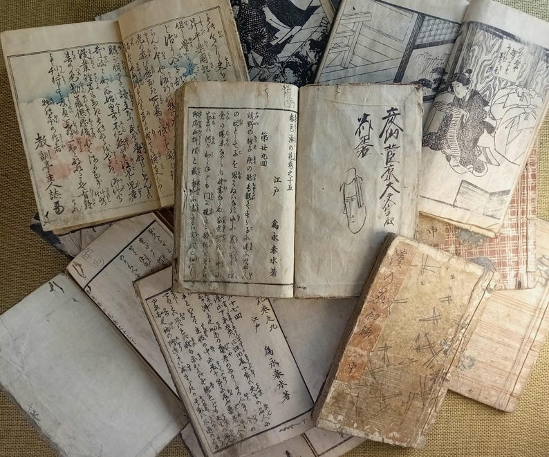 May include: A collection of antique Japanese books and documents, featuring handwritten text and illustrations. The pages are aged with visible wear and tear, showcasing the historical nature of the items. Some books are open, revealing detailed calligraphy and drawings.