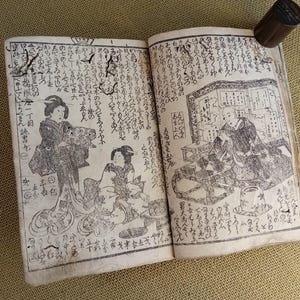 May include: An open antique book with detailed black and white illustrations and Japanese text. The illustrations depict figures in traditional clothing. A wooden stamp rests on the top right corner of the book.