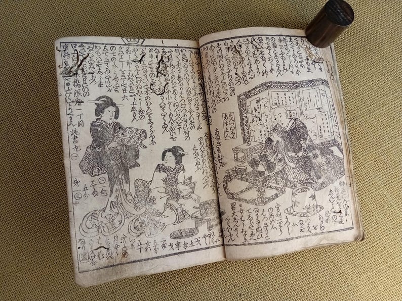 May include: An open antique book with detailed black and white illustrations and Japanese text. The illustrations depict figures in traditional clothing. A wooden stamp rests on the top right corner of the book.