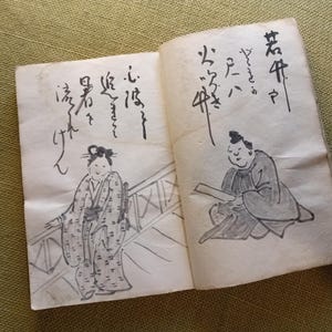 Puede incluir: Libro abierto con caligraf&iacute;a e ilustraciones japonesas. Una p&aacute;gina muestra a una mujer con kimono, la otra a un hombre leyendo. Los dibujos son en tinta negra sobre papel envejecido, lo que sugiere un art&iacute;culo vintage o antiguo.