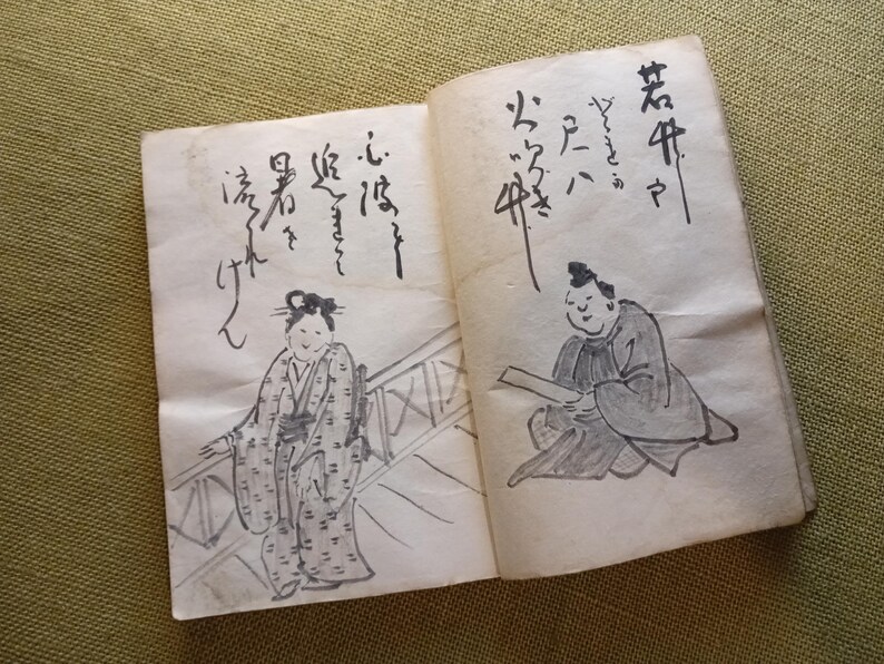 Puede incluir: Libro abierto con caligraf&iacute;a e ilustraciones japonesas. Una p&aacute;gina muestra a una mujer con kimono, la otra a un hombre leyendo. Los dibujos son en tinta negra sobre papel envejecido, lo que sugiere un art&iacute;culo vintage o antiguo.