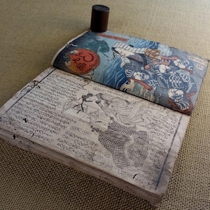 May include: An open antique book with Japanese woodblock prints. The top cover features a colourful illustration of figures and waves. The pages are filled with intricate calligraphy and drawings. A dark brown cylindrical object rests on the book.