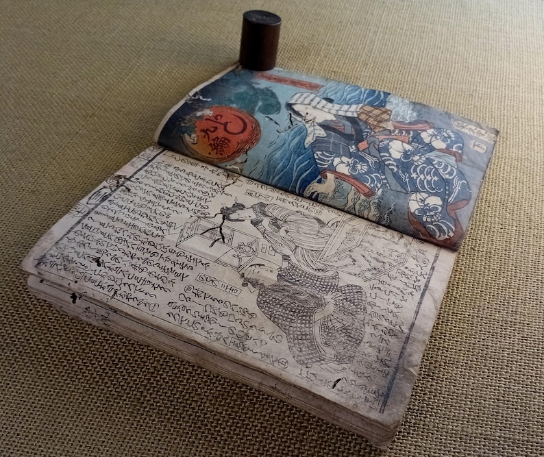 May include: An open antique book with Japanese woodblock prints. The top cover features a colourful illustration of figures and waves. The pages are filled with intricate calligraphy and drawings. A dark brown cylindrical object rests on the book.