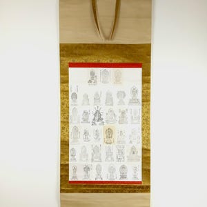May include: A traditional Japanese scroll with detailed illustrations of Buddhist figures. The scroll has a cream background with gold accents and a red border. It is suspended by tan straps. The artwork is a series of line drawings.