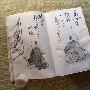 Puede incluir: Un libro abierto y envejecido con caligraf&iacute;a japonesa e ilustraciones a tinta. Los dibujos representan figuras con vestimenta tradicional y elementos arquitect&oacute;nicos. Una peque&ntilde;a piedra clara descansa cerca de la encuadernaci&oacute;n.