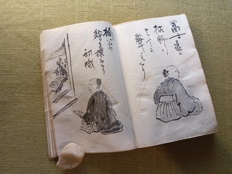 Puede incluir: Un libro abierto y envejecido con caligraf&iacute;a japonesa e ilustraciones a tinta. Los dibujos representan figuras con vestimenta tradicional y elementos arquitect&oacute;nicos. Una peque&ntilde;a piedra clara descansa cerca de la encuadernaci&oacute;n.